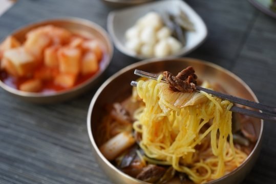 젓가락으로 든 노란 면, 뒤에 깍두기와 깐마늘
